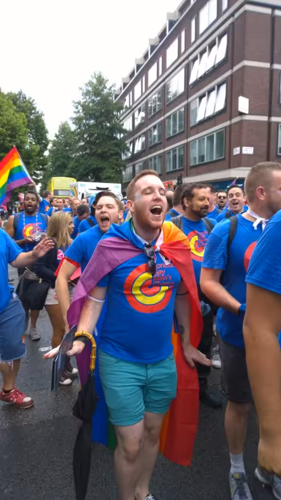 Adam at his first Pride, draped in a rainbow flag and colourful clothes, singing with the London Gay Men's Chorus