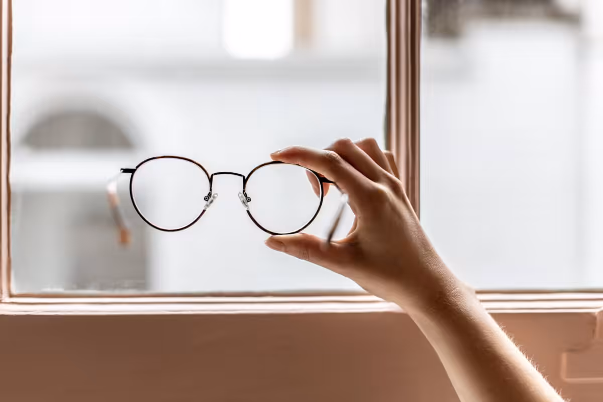 Hand holding a pair of delicate round reading glasses in front of a window