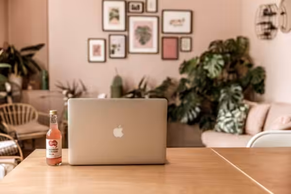 A laptop on a wooden table in front of the Studio Cotton lounge area - with black-framed artwork on the pink walls and lots of plants.