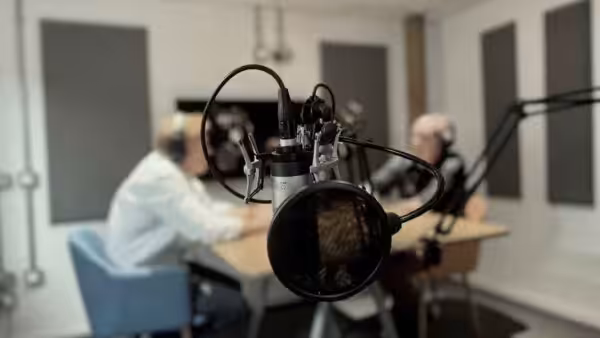 a studio mic in the foreground, with two people wearing headphones recording in the background
