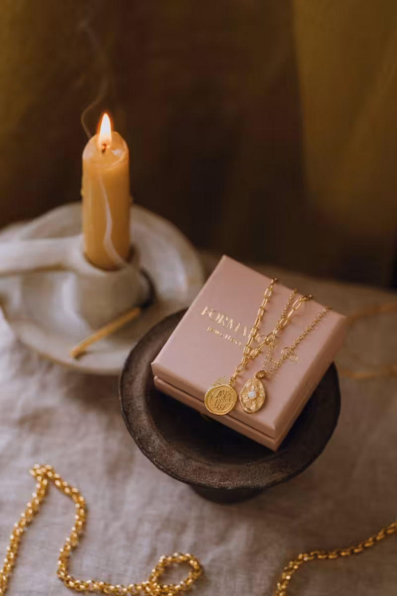 A moody shot of a lit candle and gold pendant necklaces laid over a pale pink jewellery box