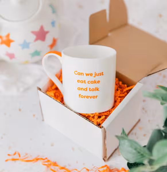 White cup in a box with orange shredded paper. Orange text on cup reads, "Can we just eat cake and talk forever?"
