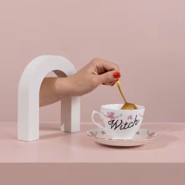 Hand reaching through white archway to stir old fashioned teacup with "witch" written on it