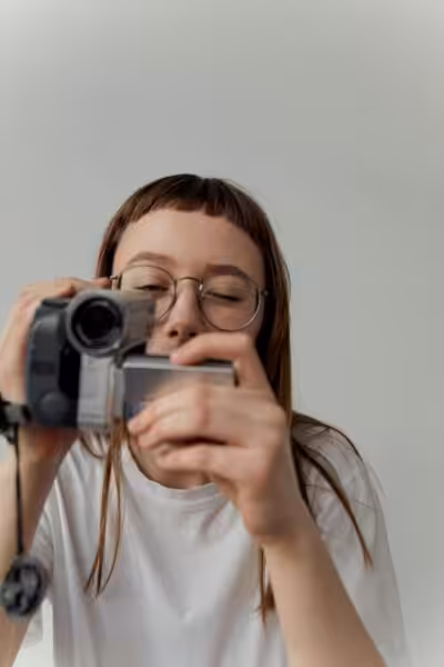 Person wearing white t-shirt and glassed holding a camcorder in front of them, looking into the viewfinder with one eye closed