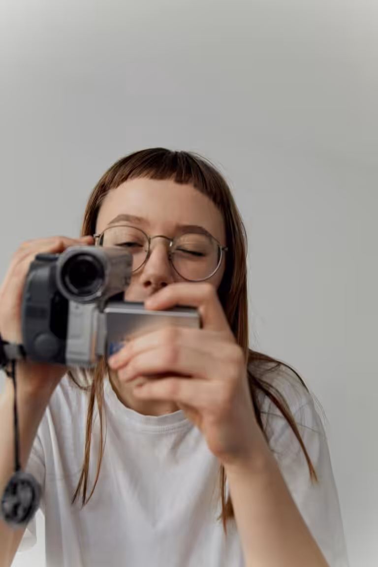 Person wearing white t-shirt and glassed holding a camcorder in front of them, looking into the viewfinder with one eye closed